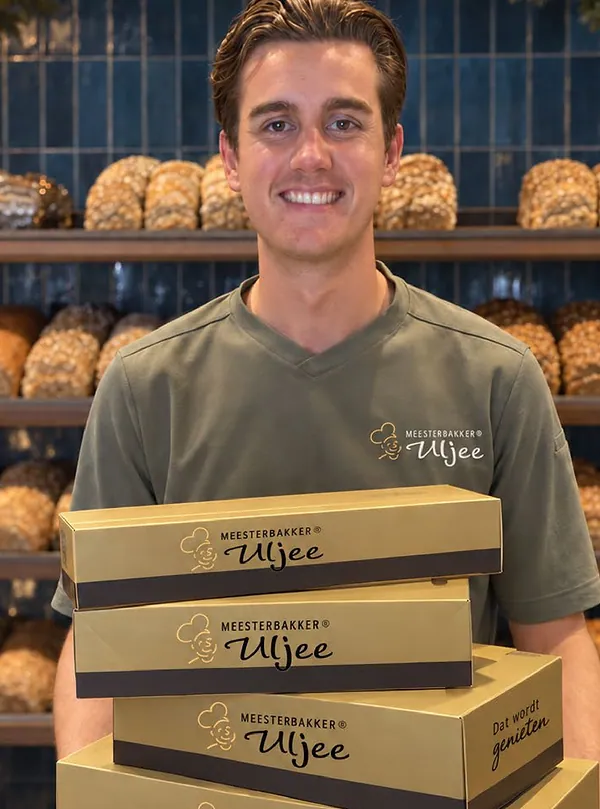 Een medewerker in een olijfgroen Uljee-shirt met vier gouden Uljee-dozen voor de blauwe tegelwand en de broodplanken