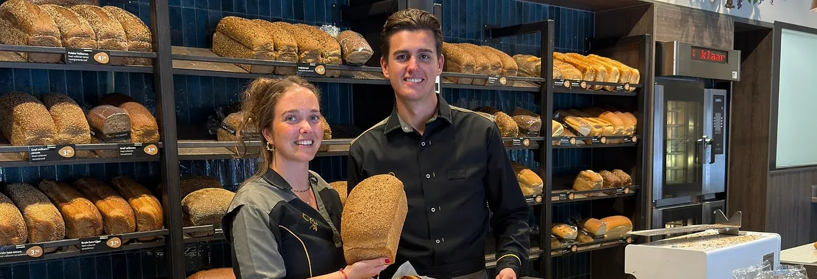 Twee medewerkers van Uljee aan de broodwand met de blauwe tegels en eiken planken, een van hen houdt een volkorenbrood vast